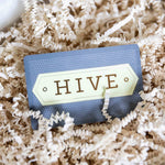 A Hive Gift Card from HIVE Home, Gift and Garden nestles in shredded beige packing paper—an ideal in-store gifting choice.