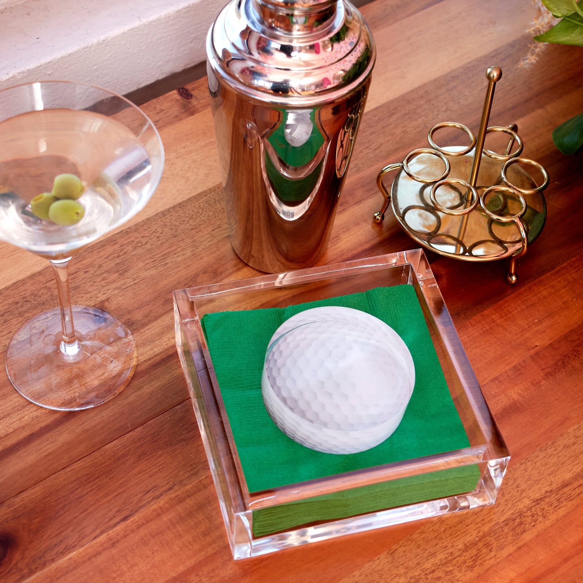A cocktail setup features a martini, metal shaker, gold holder, and the Tara Wilson Designs Lucite Cocktail Napkin Holder with Golf Ball Weight and green napkins—an ideal cocktail hour accessory on a wooden surface.