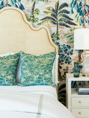 The Ingrid Bed by Oly features a beige upholstered headboard with nailhead detailing, paired with two green patterned pillows, white bedding, and a nightstand with lamp set against tropical wallpaper.