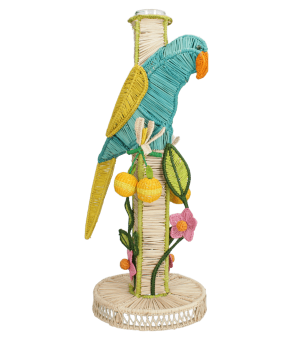 The El Pajaro Flower Vase by Mercedes Salazar features a colorful woven parrot perched on a cylindrical, parrot-adorned container, decorated with vibrant yellow fruits and pink flowers for a lively tropical touch.