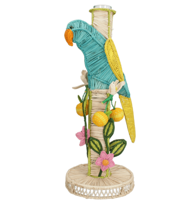 The El Pajaro Flower Vase by Mercedes Salazar features a colorful wicker parrot on a wrapped pole with yellow fruits, green leaves, and pink flowers atop a woven base, adding tropical style to your decor.
