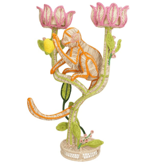The Mercedes Salazar El Mico Candle Holder features a whimsical monkey sculpture crafted from woven material, perched on a plant-inspired base with pink flowers and green leaves—combining functional décor with artistic flair.