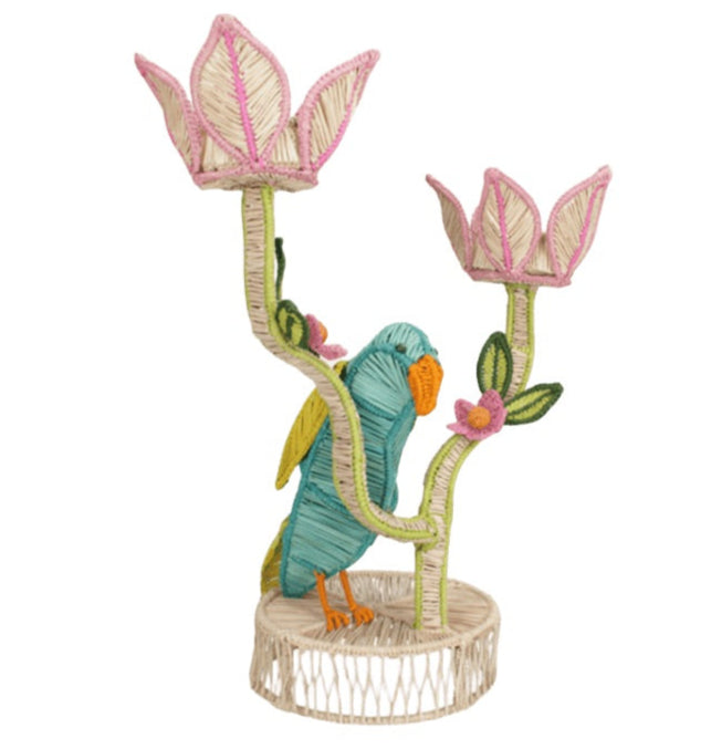 The El Loro Candle Holder by Mercedes Salazar features a parrot on a woven stand with two pink and white flower-shaped holders, adding tropical charm to any space.