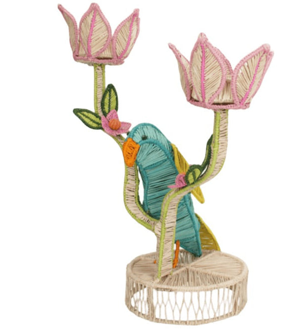 The Mercedes Salazar El Loro Candle Holder is a woven raffia centerpiece with a vibrant parrot between two pink flower-shaped holders on a round base, adding tropical cheer to any space.