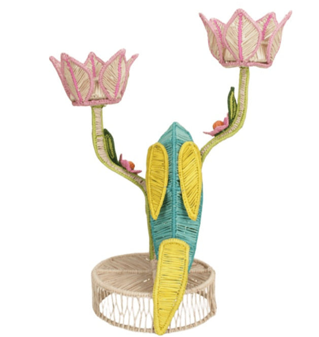The El Loro Candle Holder by Mercedes Salazar features a raffia parrot centerpiece with two pink lotus flower holders on green stems and a circular beige base, bringing tropical charm to your decor.