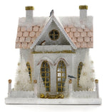 The Cody Foster Winter Cottage is a small decorative white cardboard house with glitter, two chimneys, arched windows, a frosted roof, and two white trees flanking the entrance—perfect for display or festive packaging.