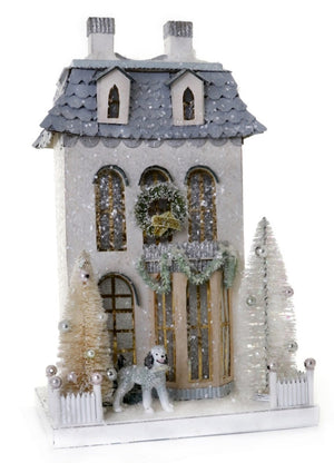 The Cody Foster White Chateau is a miniature cardboard two-story house with a snowy roof, decorated trees, a wreath above the door, white picket fences, and a small black-and-white dog in front.