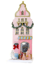 The Cody Foster Patisserie features a pink, decorative building with gold trim, garland, wreath, white dog figurine, and pink snowy Christmas trees—perfect for display or gifting in its sturdy box.