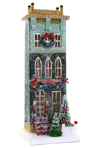 The Cody Foster Green Townhouse is a tall, rectangular green cardboard building featuring holiday wreaths, red bows, snow, small trees, and a white fence—ideal for festive packaging or decorative display.