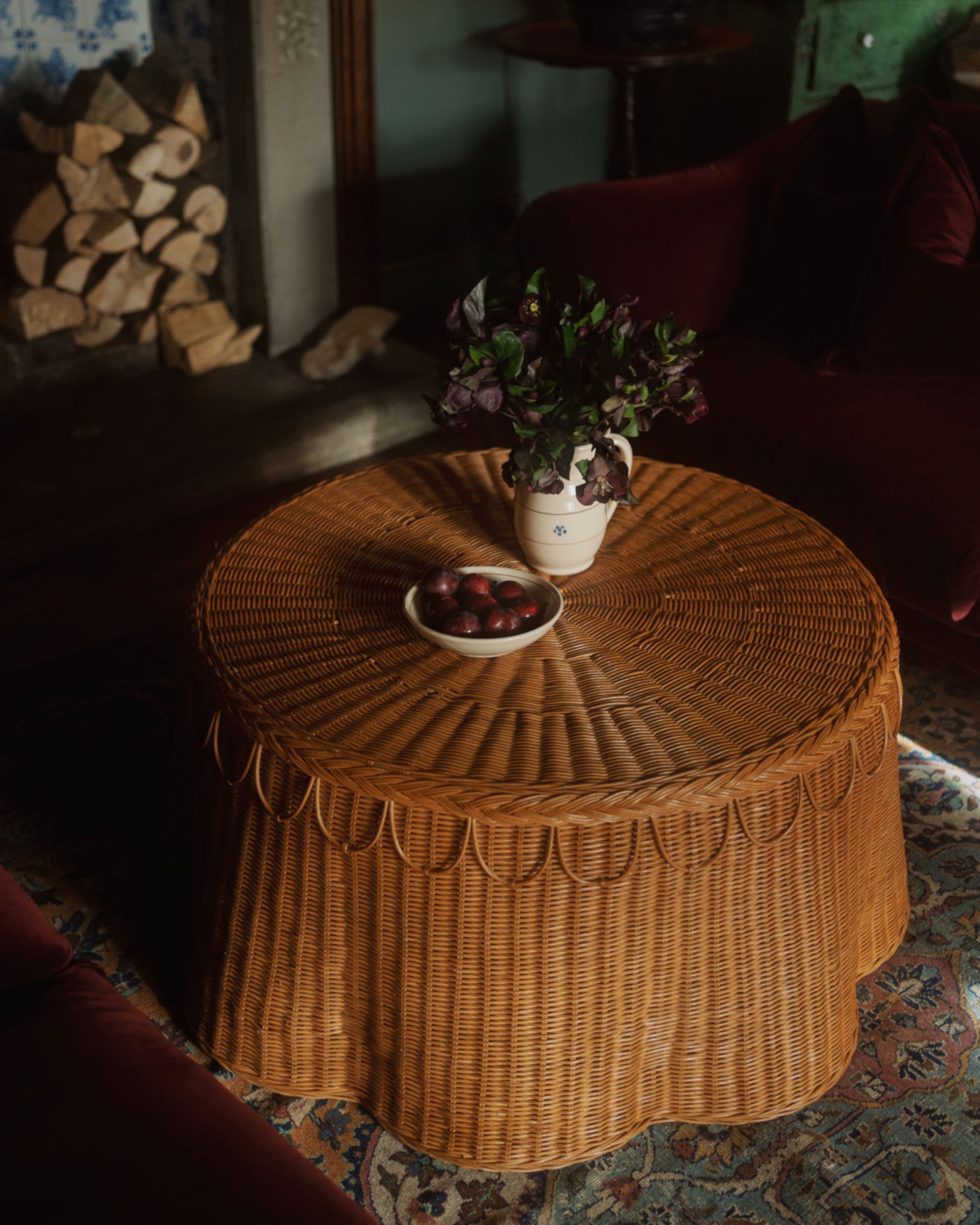 The Adeline Coffee Table by Sharland England, with its round wicker design and decorative trim, displays a vase of flowers and a bowl of plums in a cozy room featuring a fireplace and stacked firewood in the background.