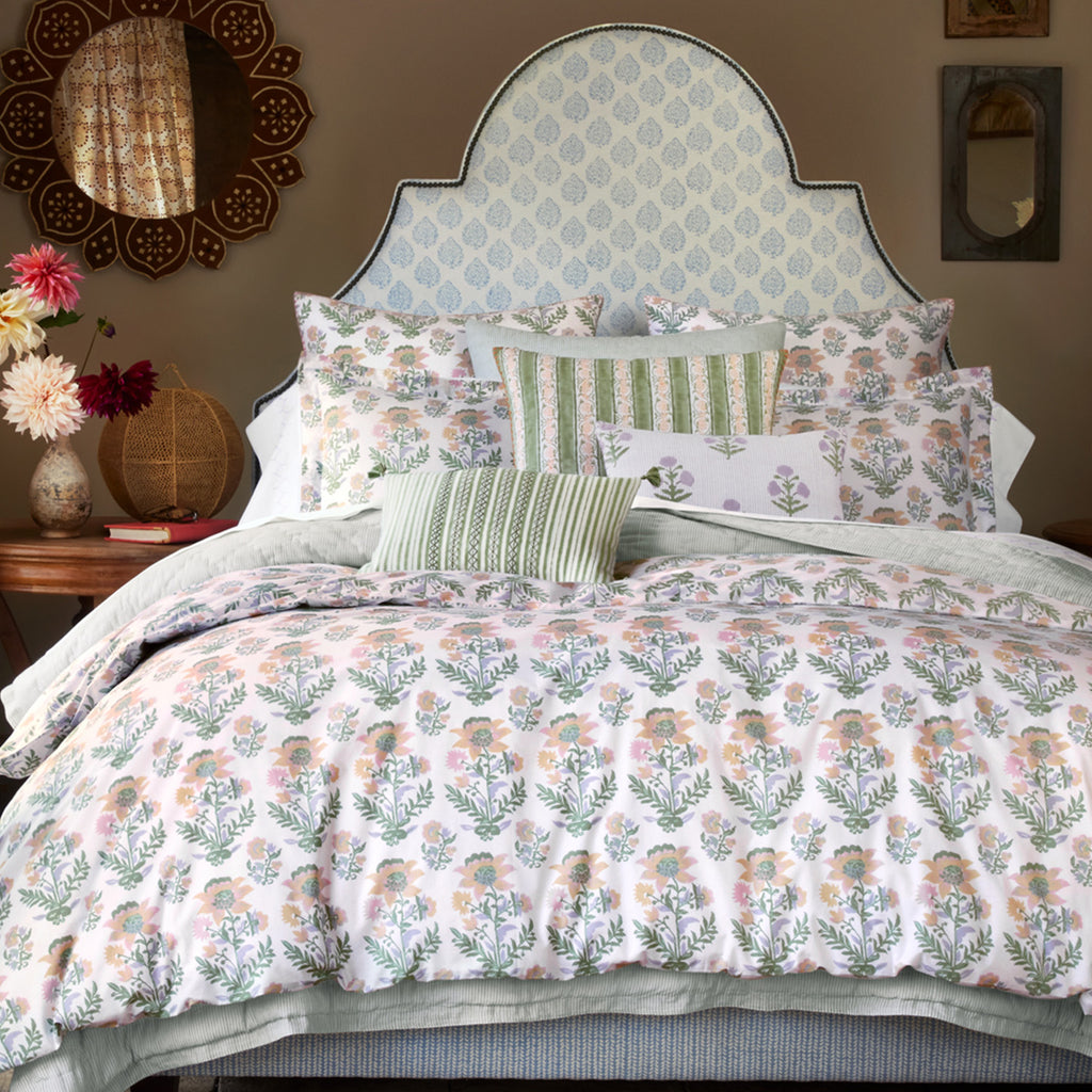 A neatly made bed features a floral comforter, assorted pillows including the John Robshaw Bhuv Sage Kidney Pillow, a pale blue upholstered headboard, and side tables with decor.