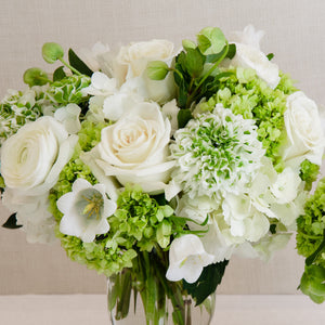 The Live Classic Floral Arrangement by Hive Floral Studio features white and light green roses, hydrangeas, and ranunculus in a clear glass vase—an elegant choice for any occasion or baby shower.