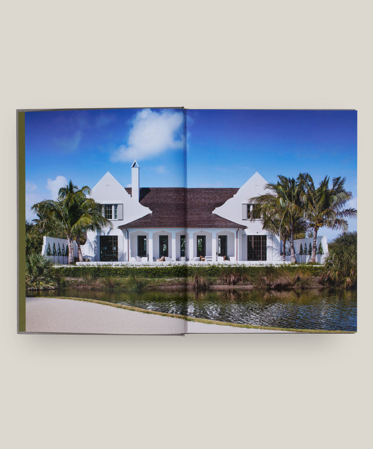 An open book, "House Love: Moor Baker Architects" by Vendome, displays a white house with a dark roof amid palm trees and a pond beneath a blue sky, highlighting Florida’s classical architecture.