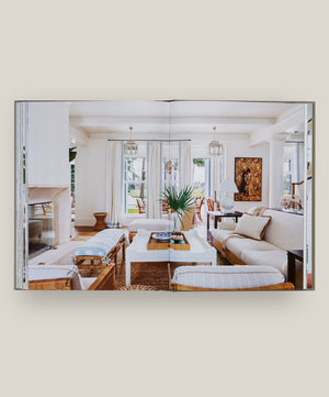 An open book, "House Love: Moor Baker Architects" by Vendome, displays a photo of a bright, modern living room with white walls, large windows, and contemporary furniture inspired by Florida architects.