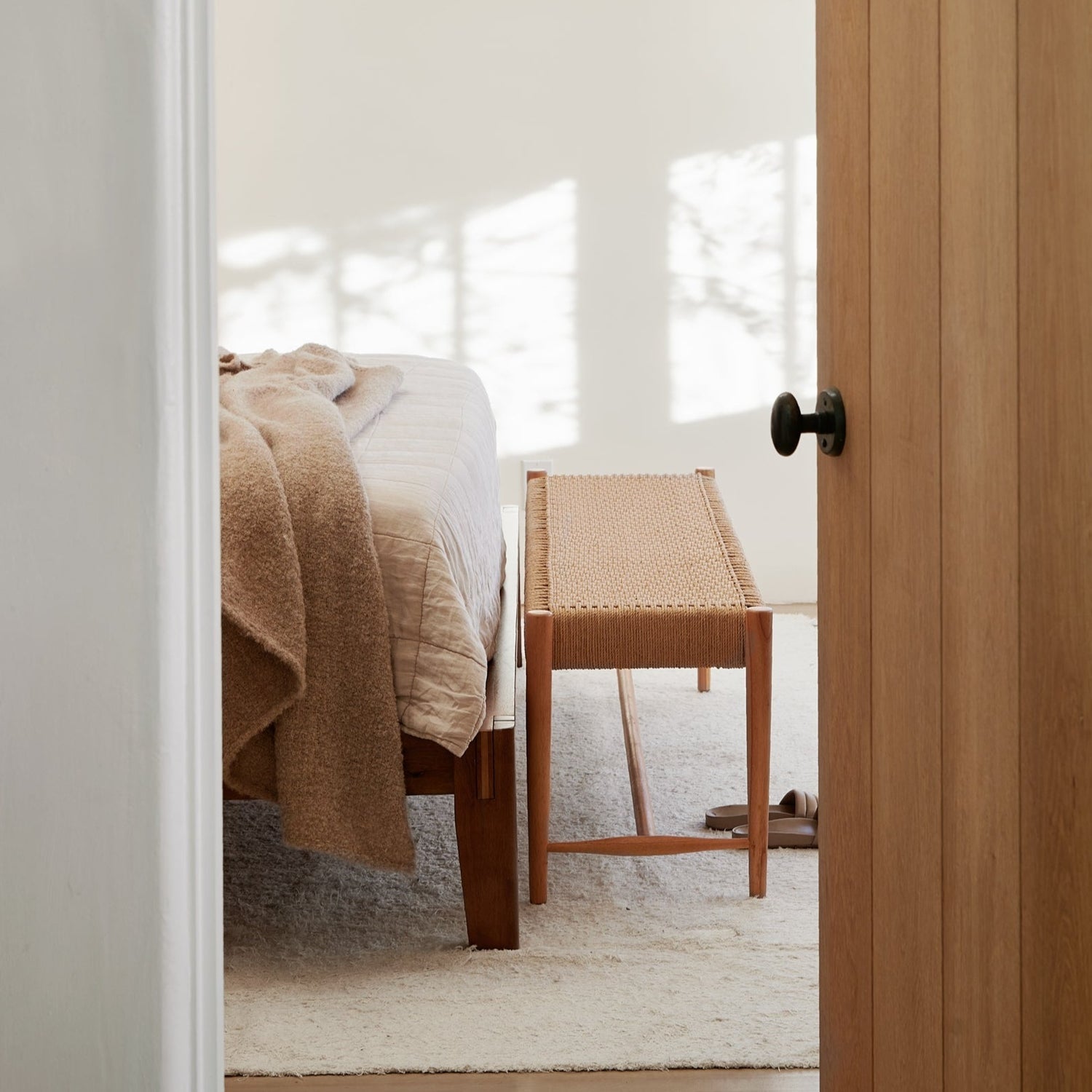 A bedroom featuring a wooden bed with beige bedding, a tan blanket, and the Selamat Ingrid Woven Bench at the foot of the bed, plus a light rug—all seen through a partially open wooden door.