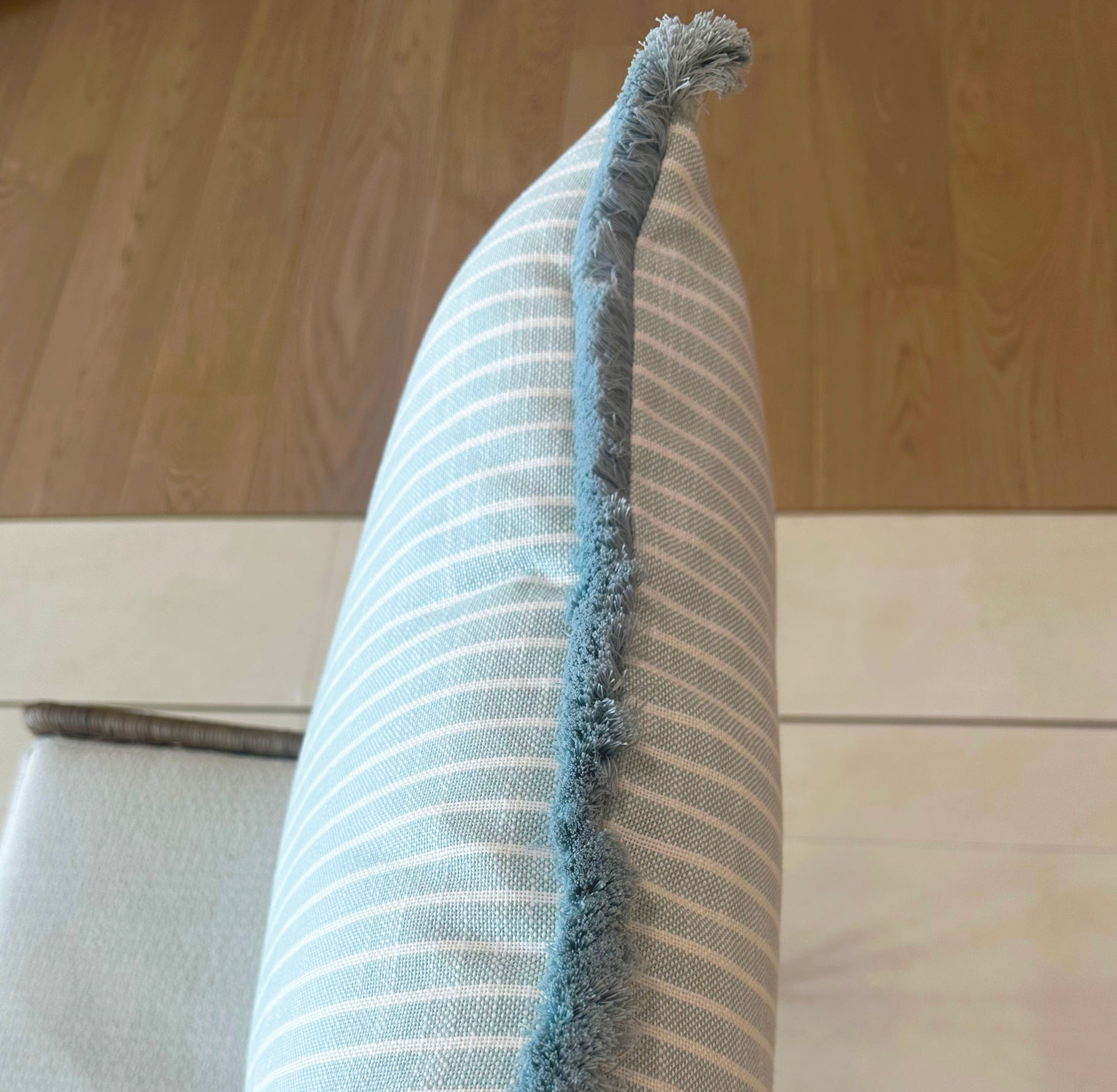 The Bayside Stripe Outdoor Pillow by Associated Design, featuring light blue and white stripes with a fringed edge, is propped upright on a chair against a backdrop of wood and tile flooring.