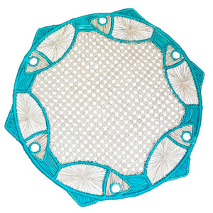 The Blue Fish Meeting Placemat by Mercedes Salazar is a round woven raffia placemat featuring turquoise edges, tropical blue fish designs, and a central lattice pattern.