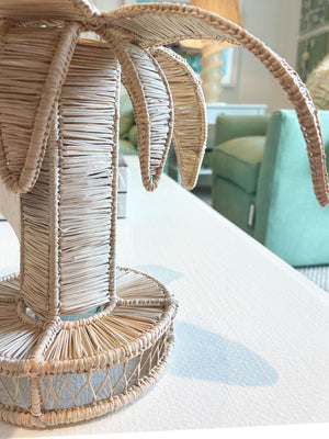 Close-up of the Mercedes Salazar Valley Flower Vase, made of woven raffia rattan on a white textured surface, with a green armchair and lamp in the background, adding a touch of tropical fun to the scene.