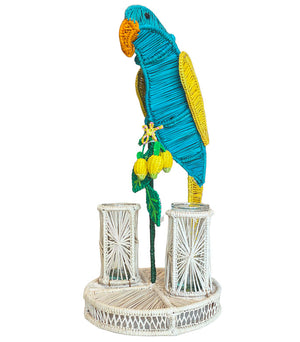 The Mercedes Salazar Lemon Tree Flower Vase features a woven wicker blue and yellow parrot perched on a white base, with decorative lemons, two small cylindrical holders, and natural raffia accents, evoking a tropical paradise.