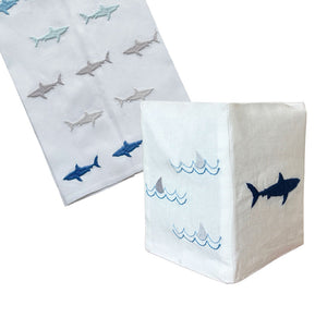 Haute Home's Shark Bath Collection features white Italian linen napkins with hand-embroidered gray and blue shark motifs and wave patterns, beautifully displayed with one folded to reveal multiple detailed designs.