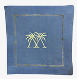 The Coastal Cocktail Napkin Set by Pink House Mustique features coastal blue linen napkins with a decorative embroidered border and two yellow palm trees stitched at the center.