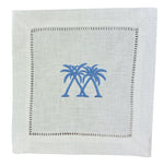The Coastal Cocktail Napkin Set by Pink House Mustique features white linen napkins with two blue embroidered palm trees at the center and a decorative hemstitch border.
