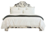 The EJ Schrader Pyne Hollyhock King Bed, featuring botanical patterns on the headboard and base, is styled with white bedding, multiple white pillows, and a dark textured throw blanket folded at the foot.