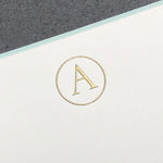Printery's Initial Card Set features a gold embossed letter "A" in a circle on white paper with an aqua border, all set against a gray background.