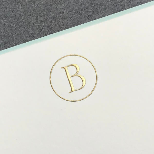 A gold initial “B” is embossed on white, encircled with elegance and set against gray—perfect for Printery’s Initial Card Set or pairing with hand-lined envelopes.