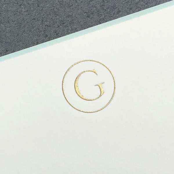 Printery’s Initial Card Set features a gold embossed “G” on white paper, framed by an aqua circle with a gray background at the top.