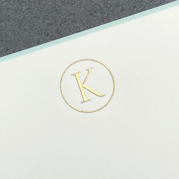 A gold initial "K" is centered in a circle on white, set diagonally by gray—capturing the classic style of Printery's Initial Card Set note cards with hand-lined envelopes.