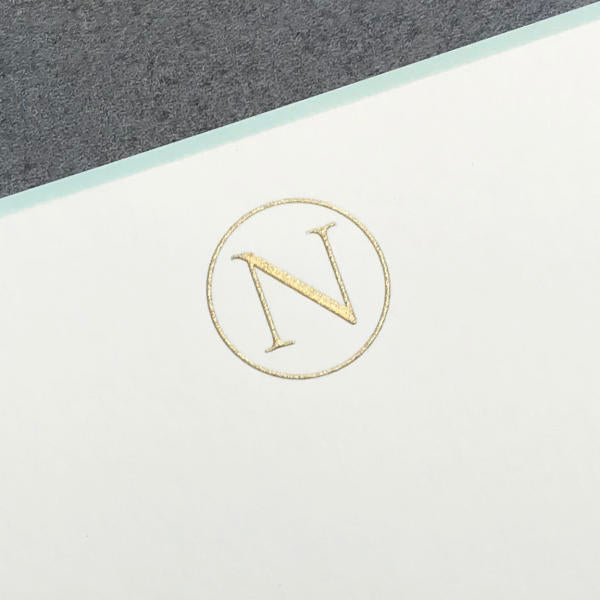 The Printery Initial Card Set features a gold letter "N" inside a circle embossed on white with an aqua border, set against gray—ideal for personalized note cards and hand-lined envelopes.