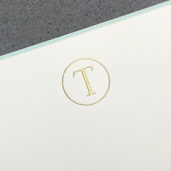 Printery's Initial Card Set features a gold “T” within a thin gold circle on white card stock against a gray background, ideal for elegant correspondence and perfect to pair with hand-lined envelopes or cards with an aqua border.
