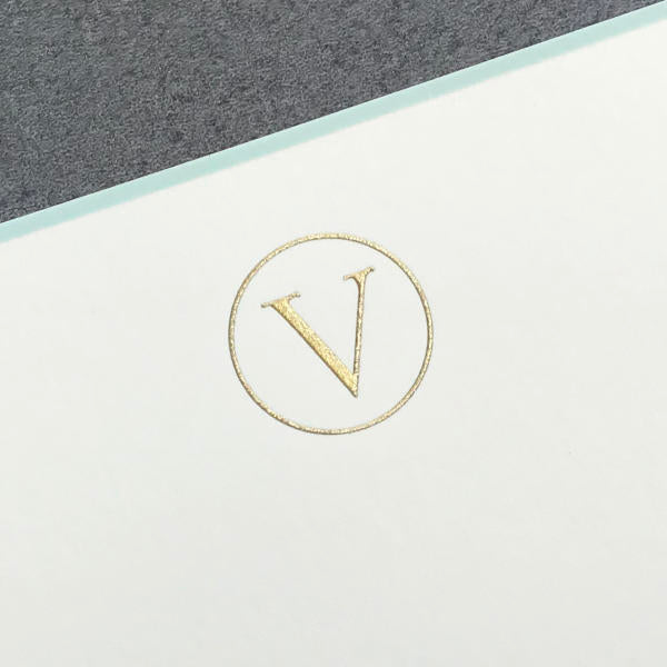 The Printery Initial Card Set features a gold "V" in a circle embossed on white paper with an aqua border, paired with hand-lined envelopes for an elegant look.