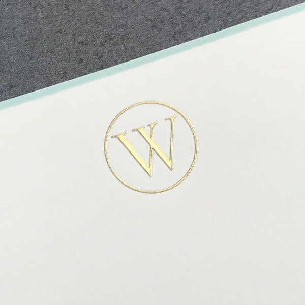 Printery’s Initial Card Set features a gold "W" embossed in a circle on white card stock with an aqua border, set against a gray background.
