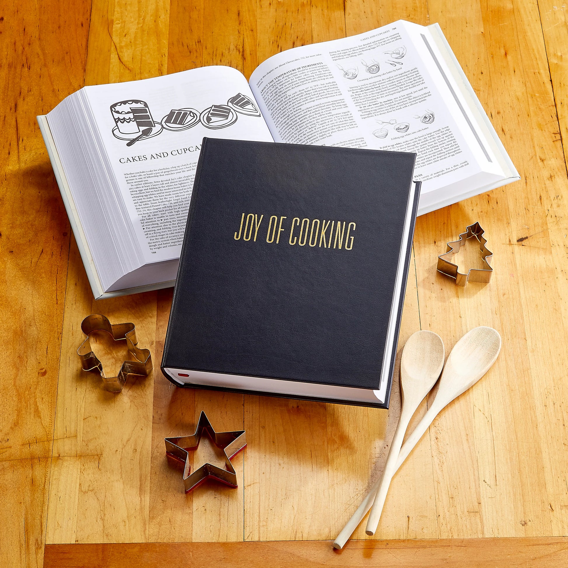 A closed Graphic Image "Joy of Cooking" cookbook rests on a wooden table, surrounded by classic recipes, wooden spoons, an open cookbook, and metal cookie cutters.
