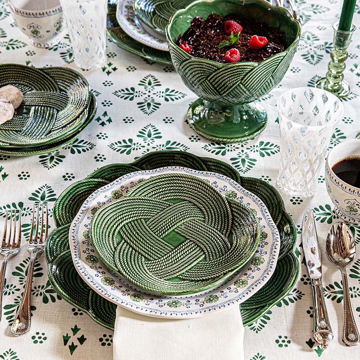 A table set with Juliska’s Le Panier Mystic Knot Basil Dinnerware and Serveware Collection, featuring patterned green and white plates and bowls, glasses, silver cutlery, and a green leaf tablecloth with ceramic bowls holding food and drinks.