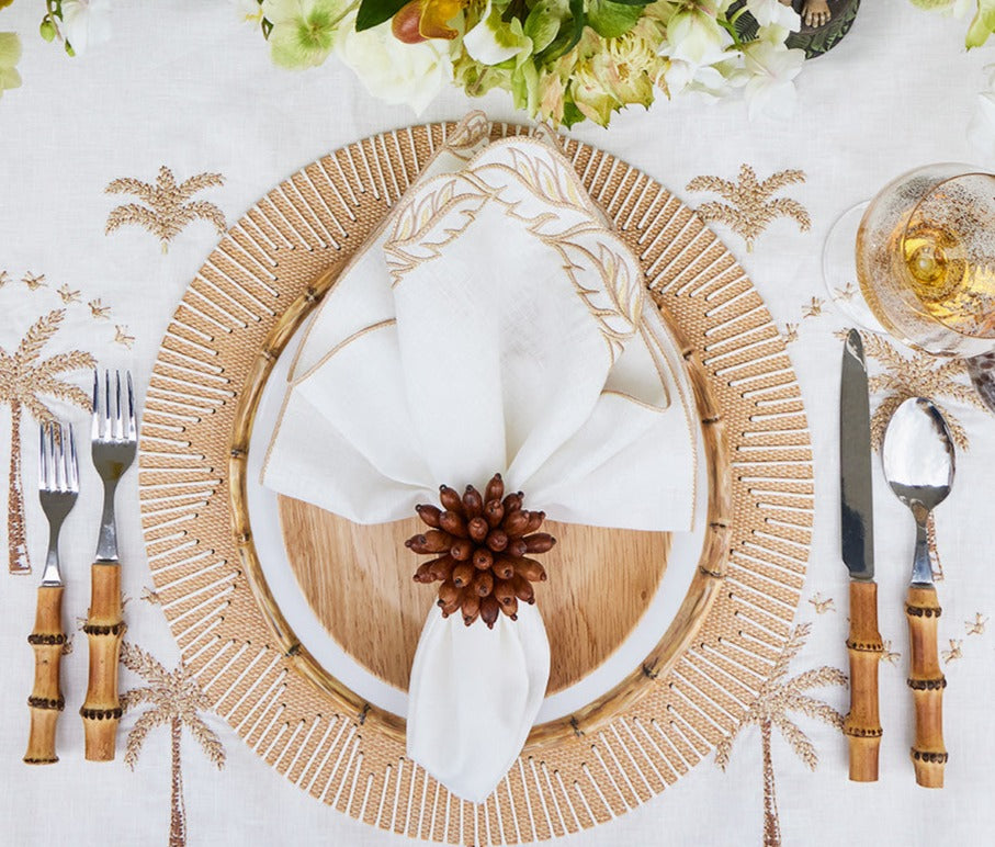 A table setting with bamboo-style flatware, wooden charger, white plate, and an embroidered napkin folded with the Kim Seybert Florette Brown Napkin Ring—ideal for outdoor gatherings and accented with palm tree motifs.