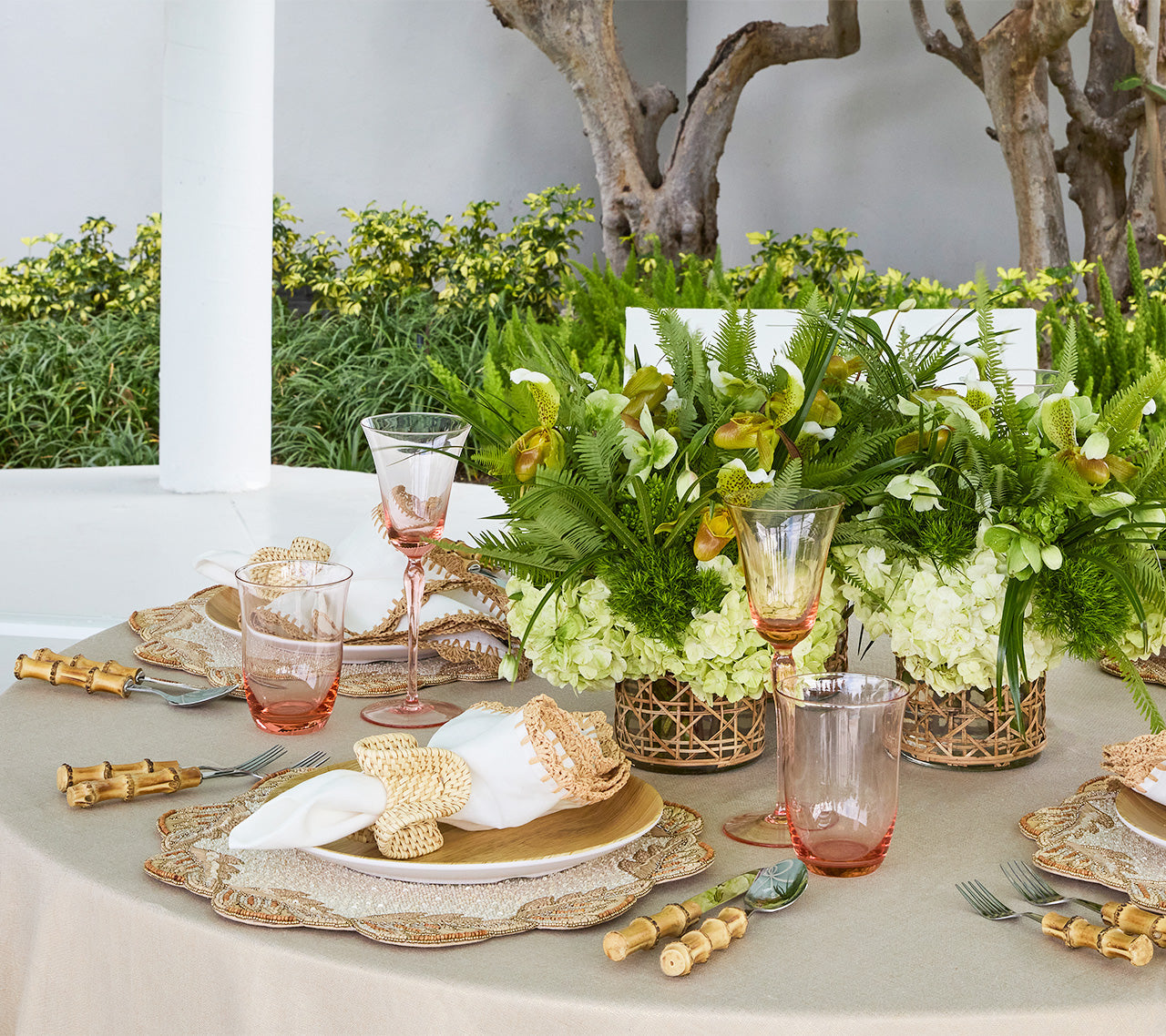Elegant outdoor table setting with gold accents, pink glassware, Kim Seybert Winding Vines Placemat, decorative napkin rings, and lush green floral centerpieces in natural tones amid greenery.