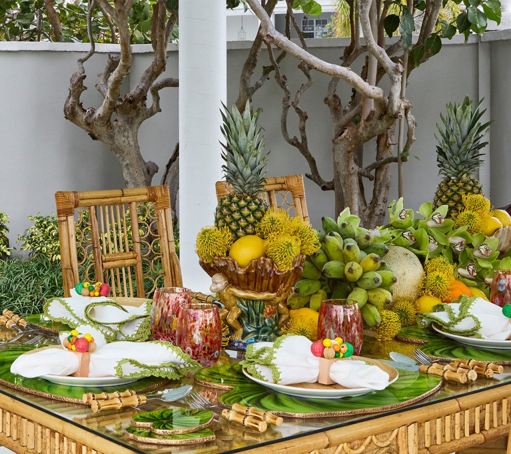 A tropical table setting features bamboo chairs, green plates on a Kim Seybert Tropicana Placemat, napkins, decorative glasses, and a centerpiece of pineapples, bananas, and yellow flowers.