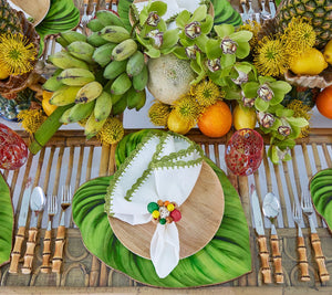 Create a tropical table setting with Kim Seybert’s Tropicana Placemat, a wooden plate, napkin, and a centerpiece featuring bananas, green orchids, and assorted fruits.