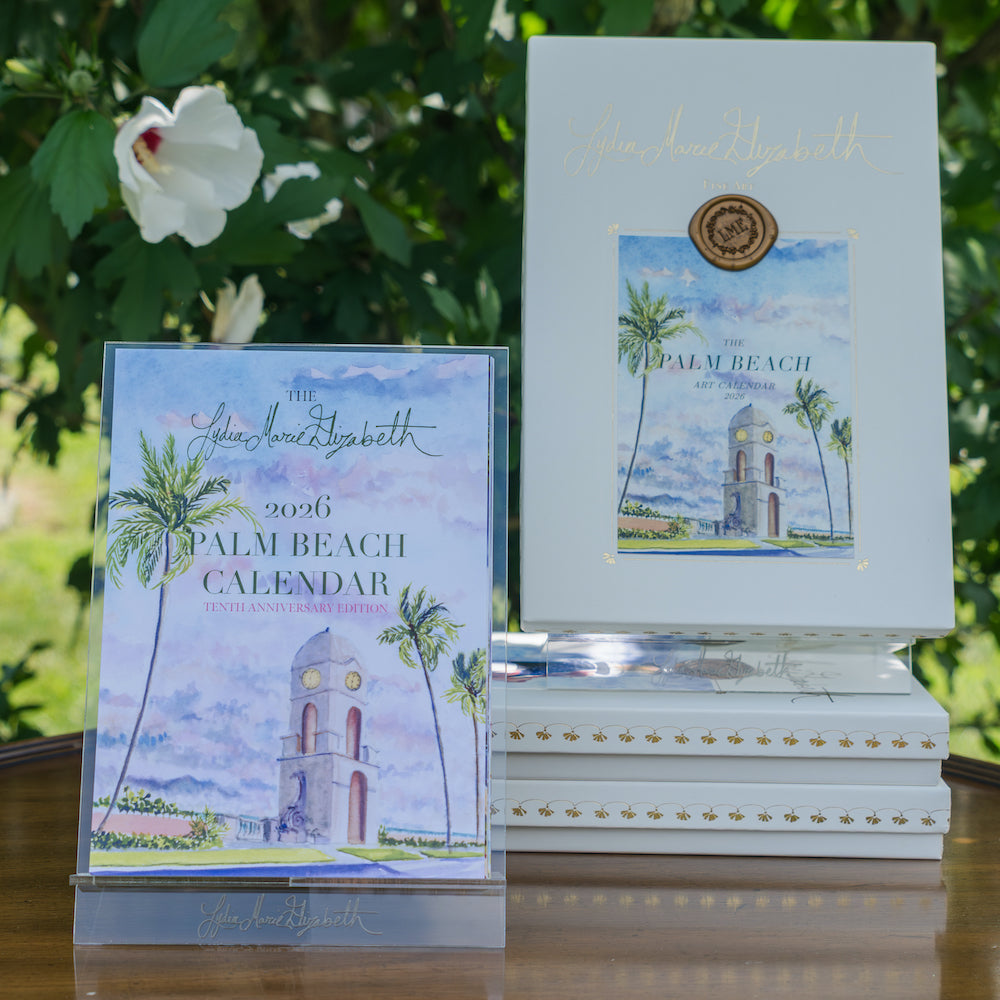 A Lydia Marie Elizabeth 2026 Palm Beach Desk Calendar with Acrylic Easel and several art-themed books sit on an outdoor table, surrounded by greenery and a white flower in the background.
