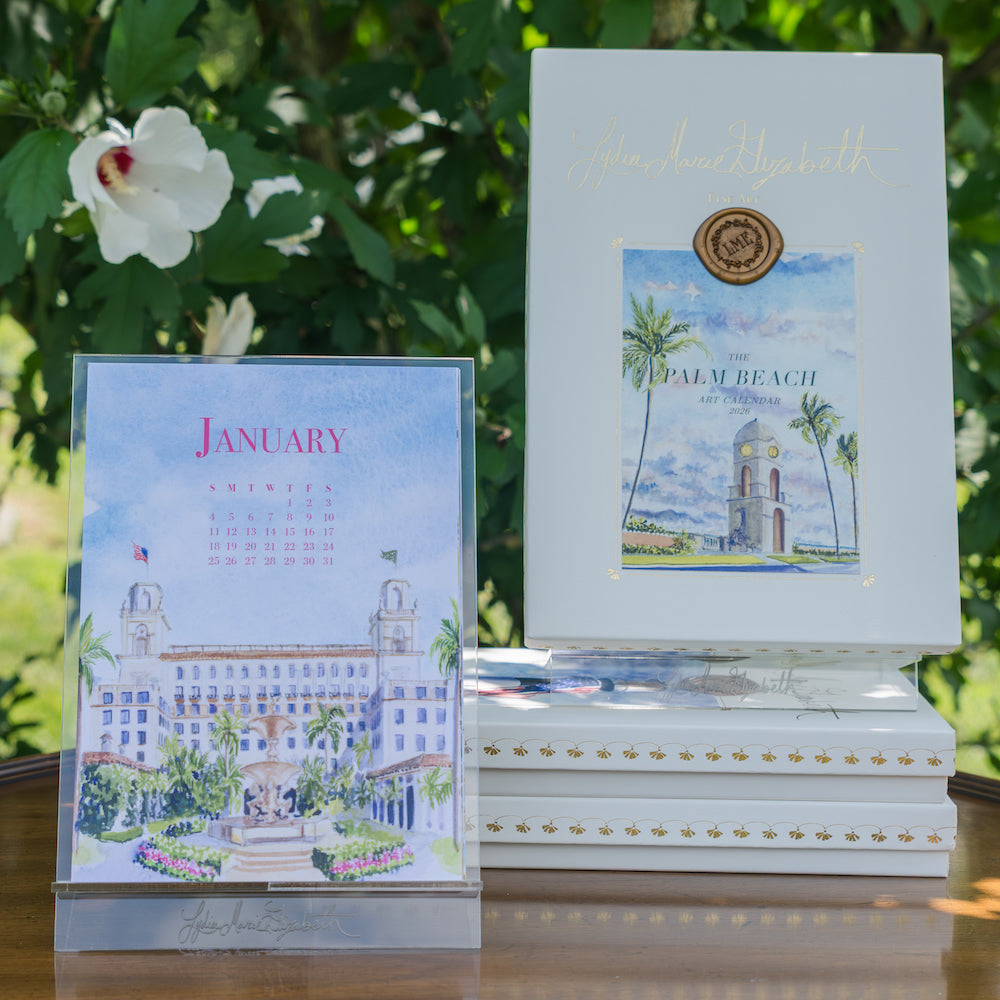 The Lydia Marie Elizabeth 2026 Palm Beach Desk Calendar with Acrylic Easel is displayed next to "The Palm Beach Art Collection" books outdoors, set against lush green foliage.
