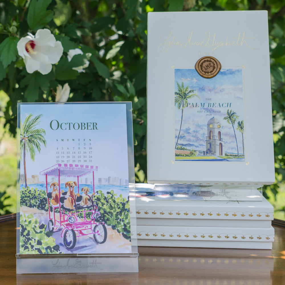 The 2026 Palm Beach Desk Calendar with Acrylic Easel by Lydia Marie Elizabeth displays its October page—dogs riding a pink bike by the beach—beside art books, lush green foliage, and white flowers.