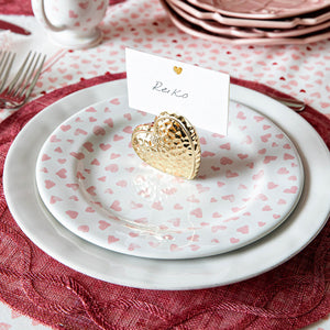 Create a romantic table with the Juliska Love You More Dessert/Salad Plate, featuring ceramic heart motifs, paired with a gold heart-shaped card holder and pink-red accents atop a lace tablecloth.