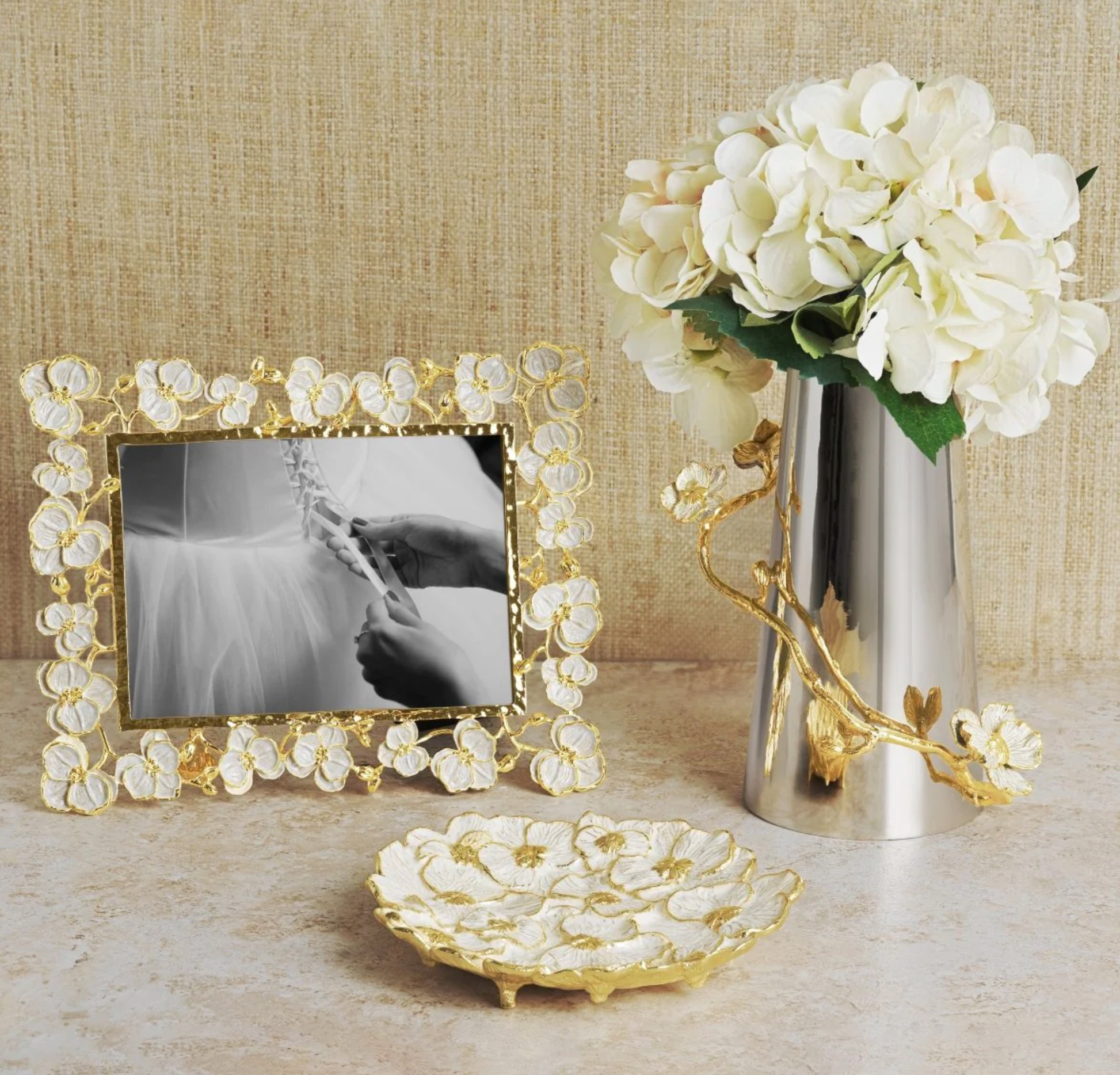 The Michael Aram Orchid Catch All, paired with a gold and white photo frame and a silver vase with white flowers, evokes subtle sensuality on a beige surface.