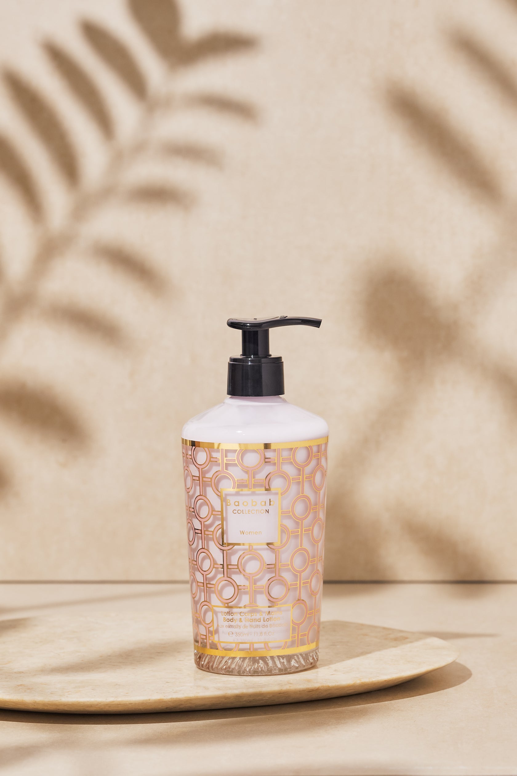 The Baobab Collection Women Body Wellness Collection by Baobab, featuring a geometric-patterned bottle with pump dispenser, rests on a round tray against a beige backdrop with leaf shadows, suggesting a subtle floral fragrance.