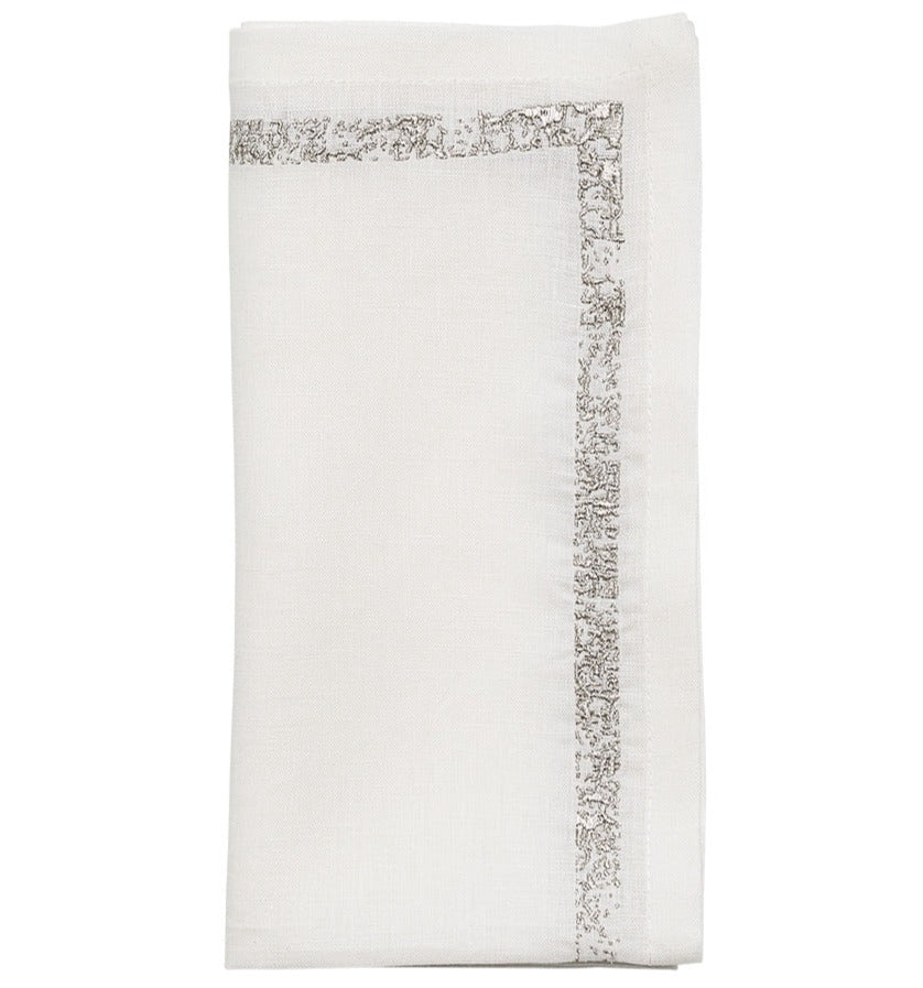 The Kim Seybert Impression Napkin Set features white linen napkins with a subtle silver embroidered border, neatly folded against a plain background.