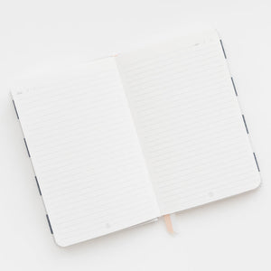 The Sugar Paper Essential Chambray Stripe Journal, featuring lined pages and a ribbon bookmark, rests on a white surface.