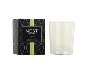 A Nest Bamboo Candle Collection candle in a white glass holder is shown next to its stylish black and green box.